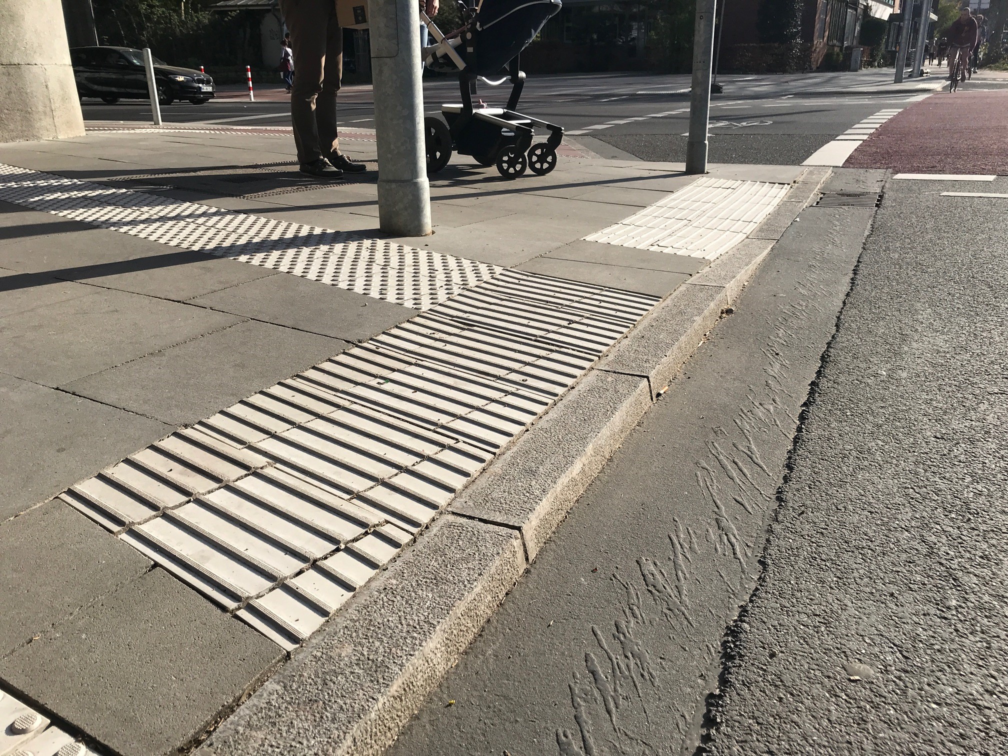 Seitlicher, flacher Blick von der Fahrbahn auf einen Bordstein: Der Bord ist im vorderen Bereich 6 cm hoch und im hinteren Bereich auf Fahrbahnniveau abgesenkt. Ein Auffindestreifen aus Noppen führt auf das Richtungsfeld am hohen Bord, der Ampelmast steht neben dem Auffindestreifen. Die Nullabsenkung ist mit einem Sperrfeld abgesichert.