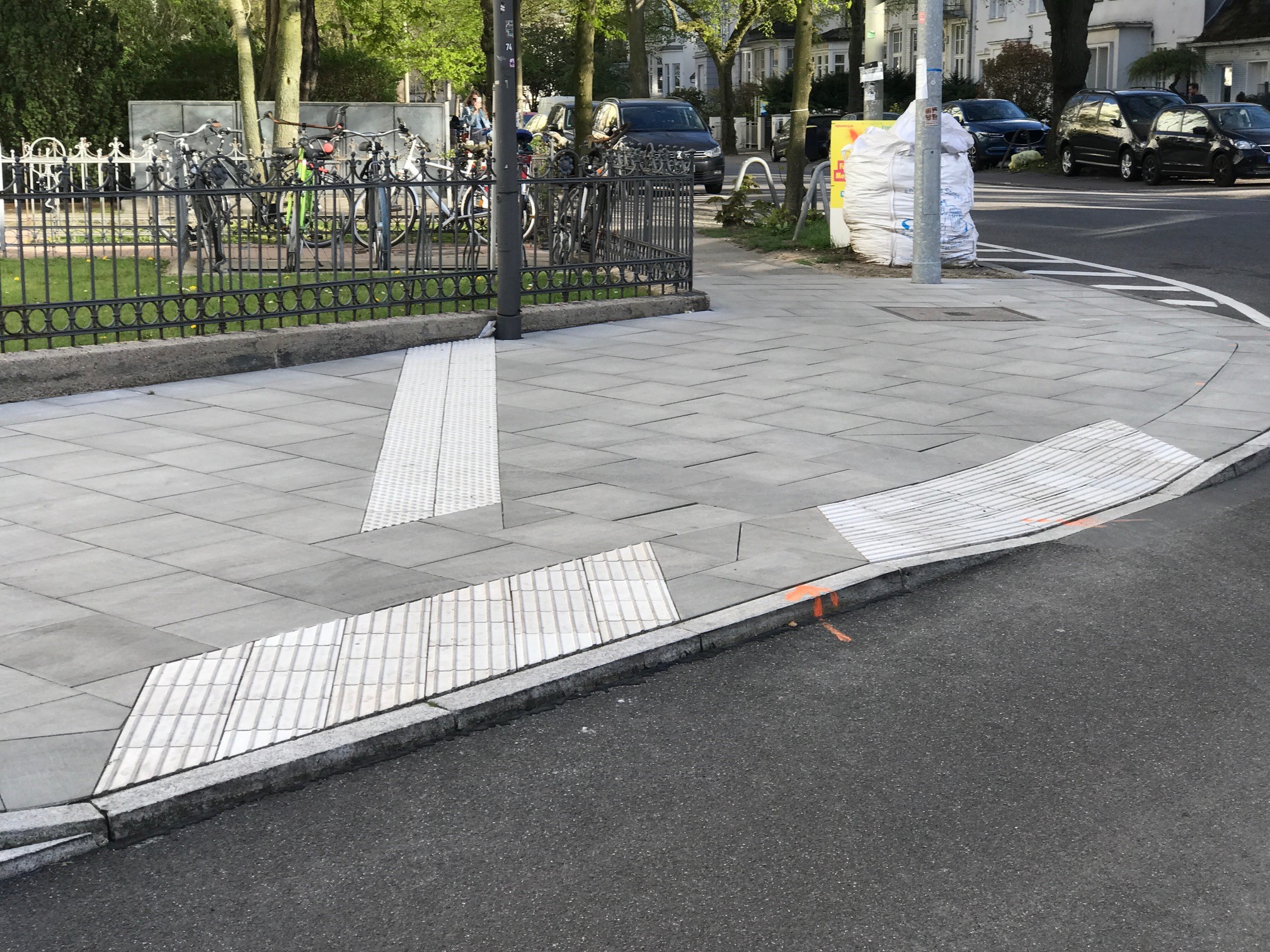 Blick von der Fahrbahn auf eine ungesicherte Querung:  Der Bordstein im linken Bereich ist 6 cm hoch und im rechten Bereich auf Fahrbahnniveau abgesenkt. Ein Auffindestreifen aus Noppen führt auf das Richtungsfeld am hohen Bord, hört jedoch etwa 90 cm davor auf. Die Nullabsenkung ist mit einem Sperrfeld abgesichert.